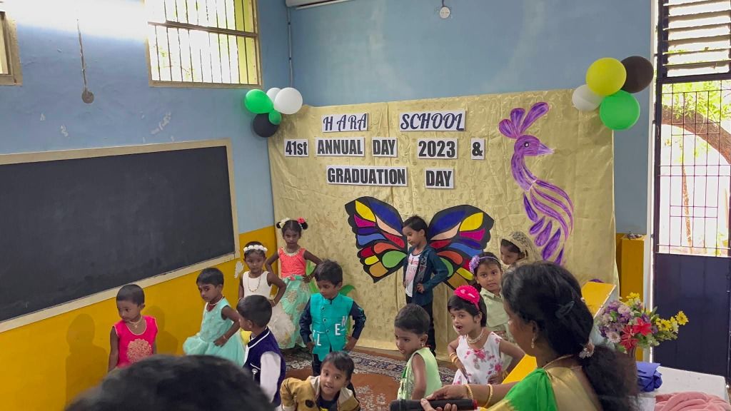 Young children in party clothes on a classroom stage in front of a butterfly-wing cut-out and a purple peacock, with a teacher holding a microphone