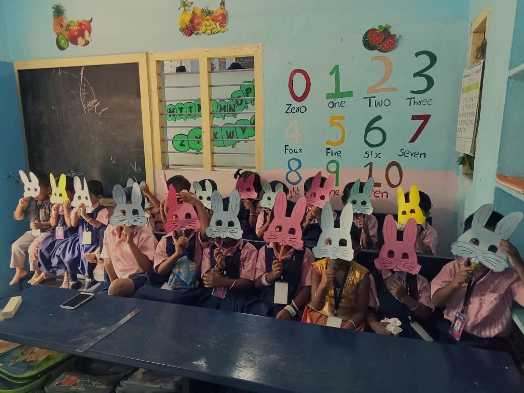 A row of children in pink-and-navy uniforms holding colourful paper bunny masks in front of their faces, beside a painted numbers wall